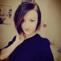 Portret młodej kobiety z krótkimi, brązowymi włosami z rudymi refleksami, uśmiechającej się do kamery, w ciemnej bluzce, z dłonią na szyi.