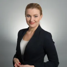 Portret uśmiechniętej kobiety w ciemnej marynarce i jasnej bluzce, z włosami upiętymi w koński ogon i delikatnym makijażem, na szarym tle.