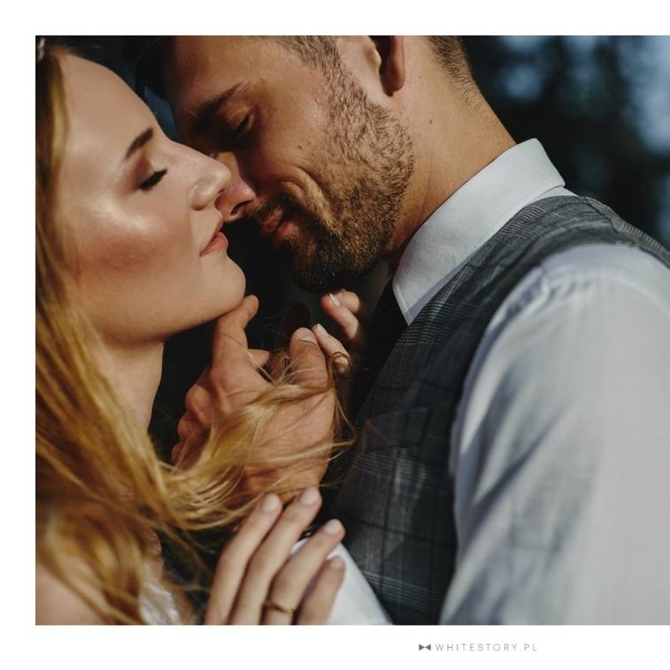 Portret narzeczeństwa w zbliżeniu: panna młoda z delikatnym, rozświetlonym upiększeniem skóry i zamkniętymi oczami, dotykana przez pana młodego w eleganckiej kamizelce, w tle zieleń.