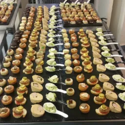 Różnorodne, starannie przygotowane finger food na ciemnych, łupkowych tacach, część z nich podana na białych i czarnych łyżeczkach; widoczne tartaletki z mięsem, pasztetem i piklami, mini kanapki...