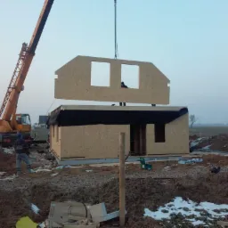 Montaż domu modułowego: dźwig umieszcza prefabrykowany element ściany z otworami okiennymi na konstrukcji, widoczny pracownik w otworze okiennym, teren budowy z resztkami śniegu.