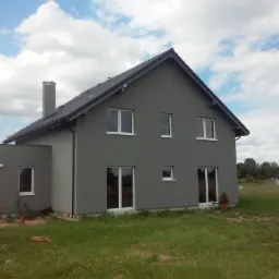 Dwukondygnacyjny dom jednorodzinny w szarej elewacji z ciemnym dachem, widoczny z zewnątrz w pochmurny dzień, z kominem i małym drzewkiem w doniczce obok okna.