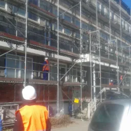 Pracownicy w pomarańczowych kamizelkach na rusztowaniu przy budynku wielopiętrowym, przygotowującym się do prac elewacyjnych, okna zabezpieczone folią.