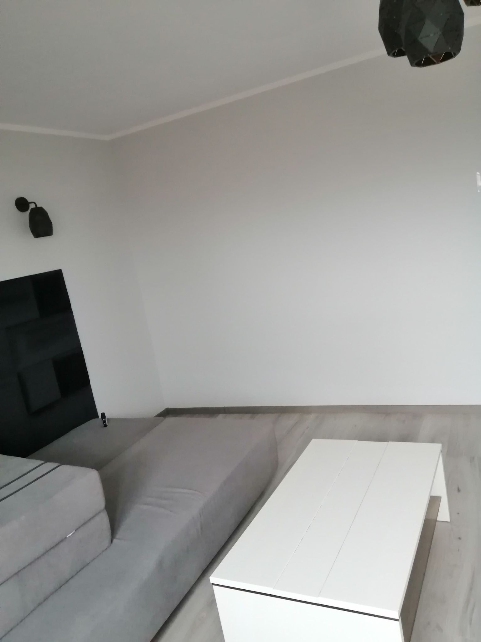 Minimalistyczne wnętrze z szarą sofą modułową i białym stolikiem kawowym na tle gładkiej, białej ściany z zamontowanym oświetleniem punktowym i geometryczną lampą wiszącą.