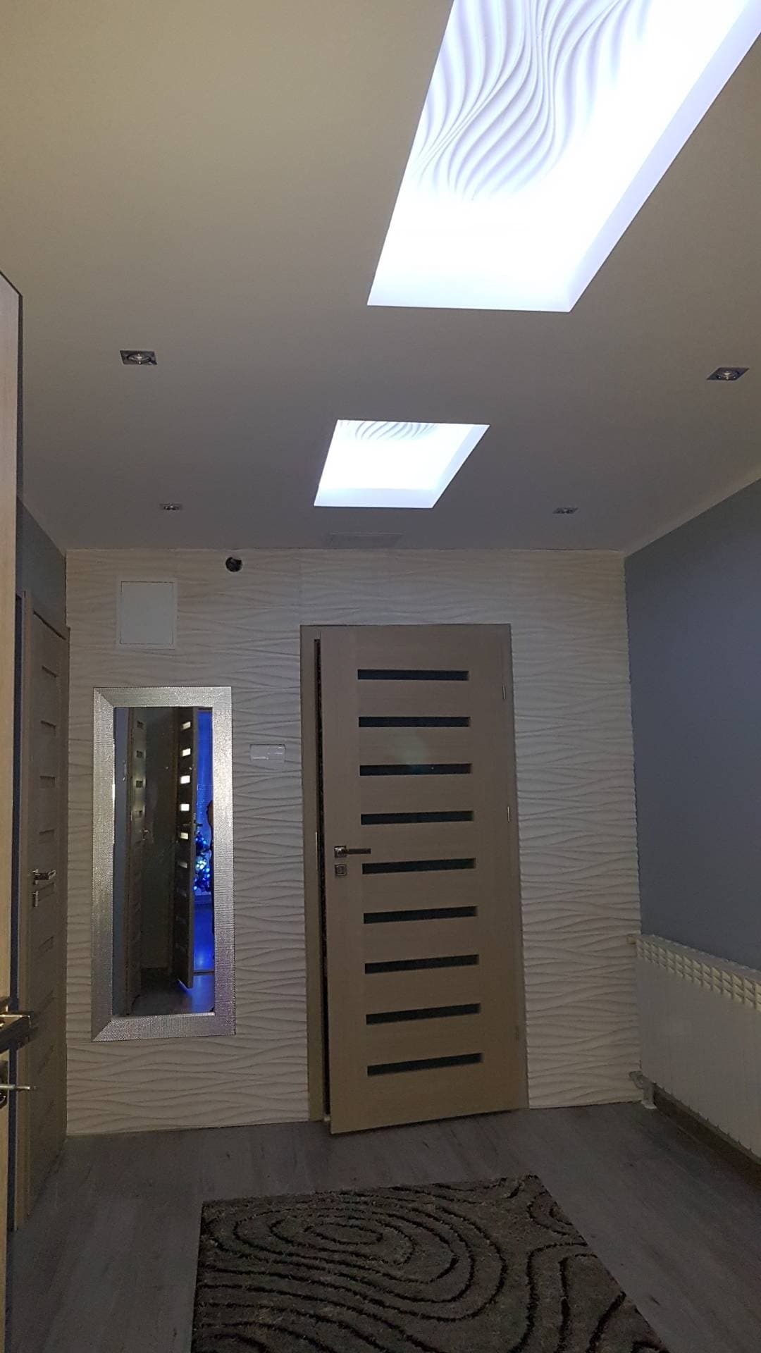 Korytarz z dwoma podświetlanymi sufitami o wzorze falistym, lustrem w srebrnej ramie i drzwiami z poziomymi przeszkleniami.