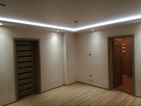 Wykończone wnętrze pokoju z dwiema parami drzwi, podwieszanym sufitem z oświetleniem LED i parkietem. Widoczne gniazdka elektryczne na ścianie.