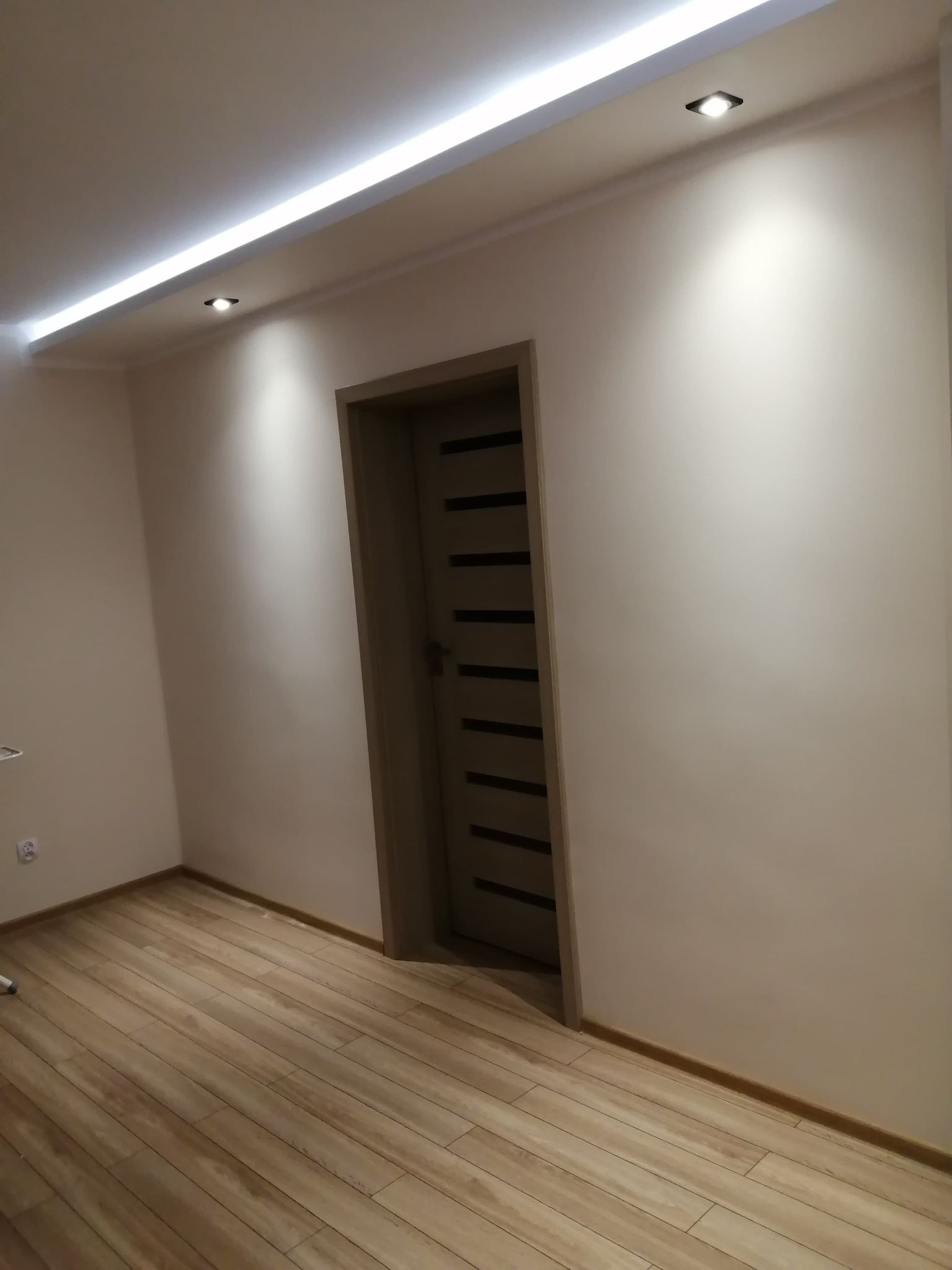 Jasne wnętrze z drewnopodobną podłogą, oświetleniem LED w suficie podwieszanym i nowoczesnymi drzwiami z poziomymi wstawkami.