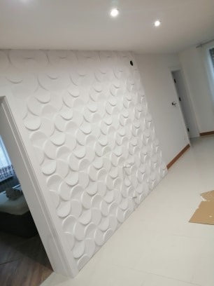 Ściana wykończona białymi panelami 3D z motywem półokręgów, widoczne oświetlenie sufitowe i fragment podłogi z listwą przypodłogową.