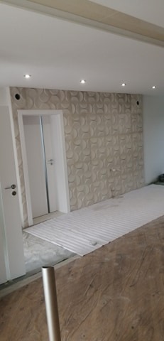 Wnętrze w trakcie remontu: ściana z dekoracyjną trójwymiarową fakturą, widoczne punkty oświetleniowe w suficie, zabezpieczona podłoga, białe drzwi.