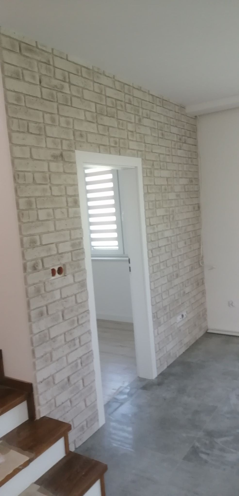 Ściana wykończona dekoracyjną cegłą w odcieniach bieli i szarości, z widocznym gniazdkiem elektrycznym i fragmentem drewnianych schodów, prowadząca do pomieszczenia z żaluzjami okiennymi.