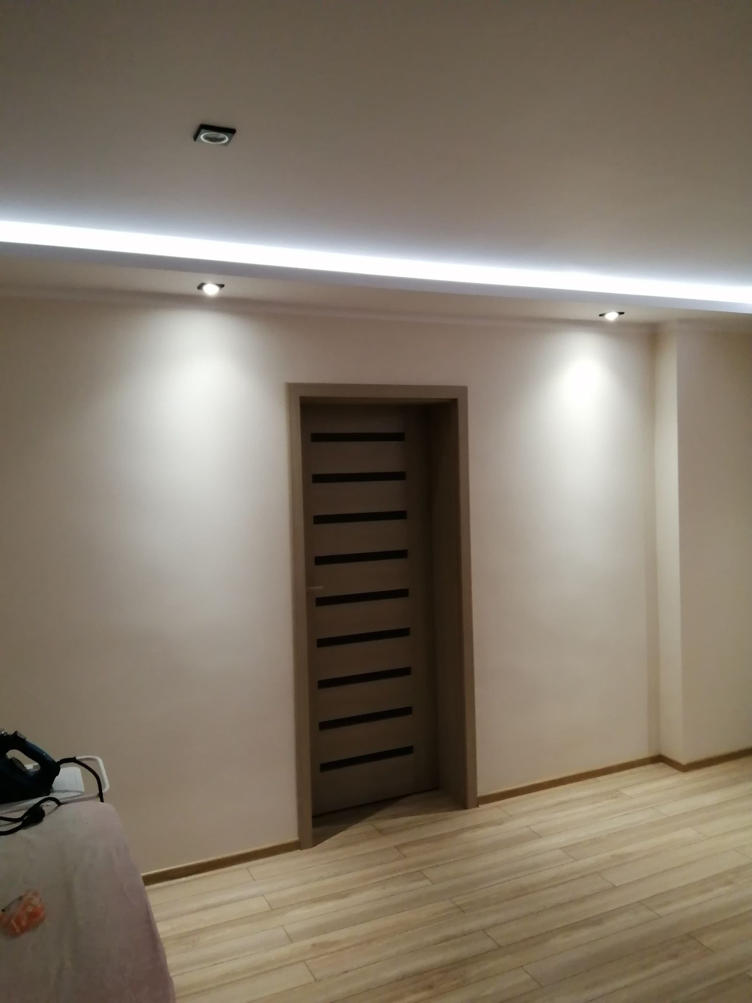 Nowoczesne wnętrze z oświetleniem LED w suficie, punktowe lampy sufitowe i drewniane panele podłogowe, widoczny fragment deski do prasowania z żelazkiem.