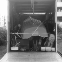 Wnętrze ciężarówki załadowane różnorodnymi przedmiotami, w tym odwróconą łodzią zabezpieczoną pasami, widok z zewnątrz z otwartą klapą, czarno-białe zdjęcie.