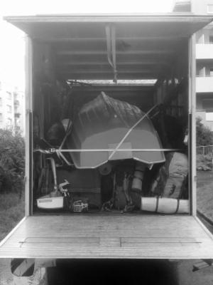 Wnętrze ciężarówki załadowane różnorodnymi przedmiotami, w tym odwróconą łodzią zabezpieczoną pasami, widok z zewnątrz z otwartą klapą, czarno-białe zdjęcie.