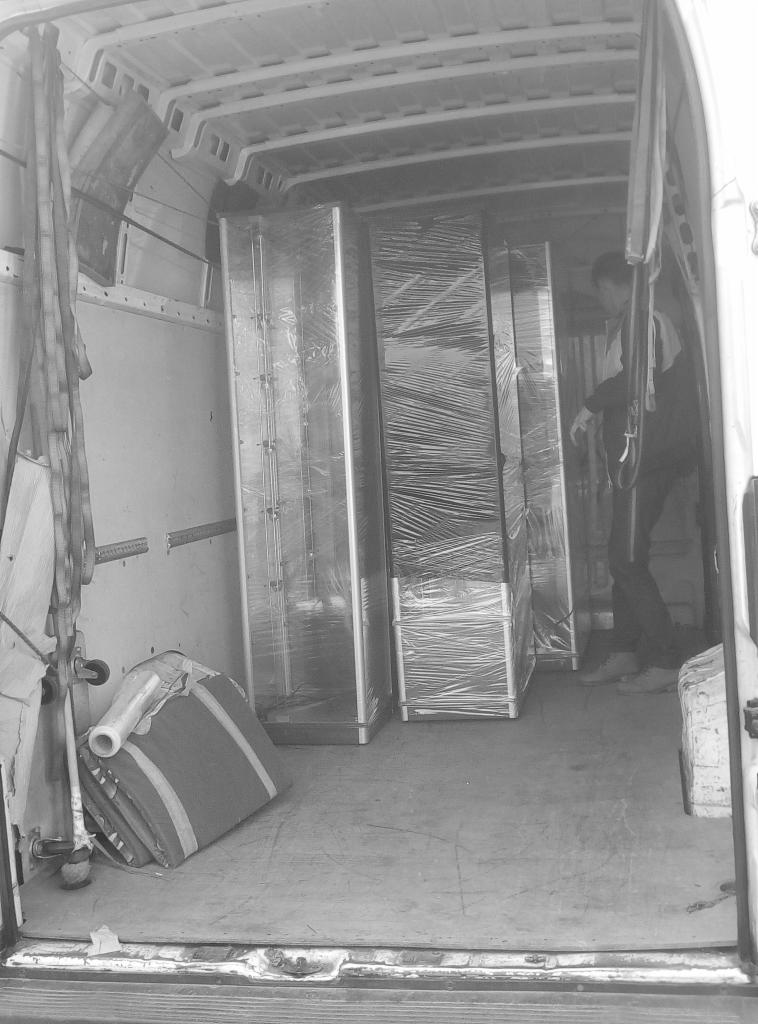 Wnętrze furgonetki z załadowanymi szafkami owiniętymi folią stretch, zabezpieczone pasami transportowymi, widoczny pracownik w tle.