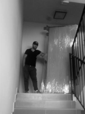 Mężczyzna w czarnej koszulce i czapce opiera się o zabezpieczony folią mebel na schodach w klatce schodowej.