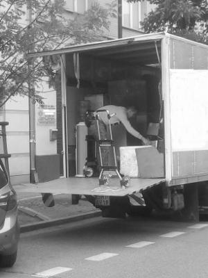 Czarno-białe zdjęcie przedstawia pracownika na drabinie wewnątrz otwartej ciężarówki, układającego kartony i zabezpieczone meble; widoczny fragment ulicy i zaparkowany samochód.
