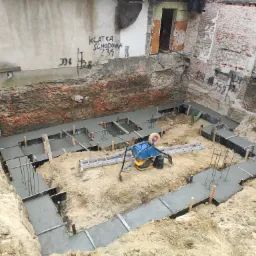 Wylane betonowe ławy fundamentowe w wykopie, z widocznymi szalunkami, wystającymi prętami zbrojeniowymi, ubijarką wibracyjną i drabiną, na tle starego, częściowo zniszczonego budynku z cegły...