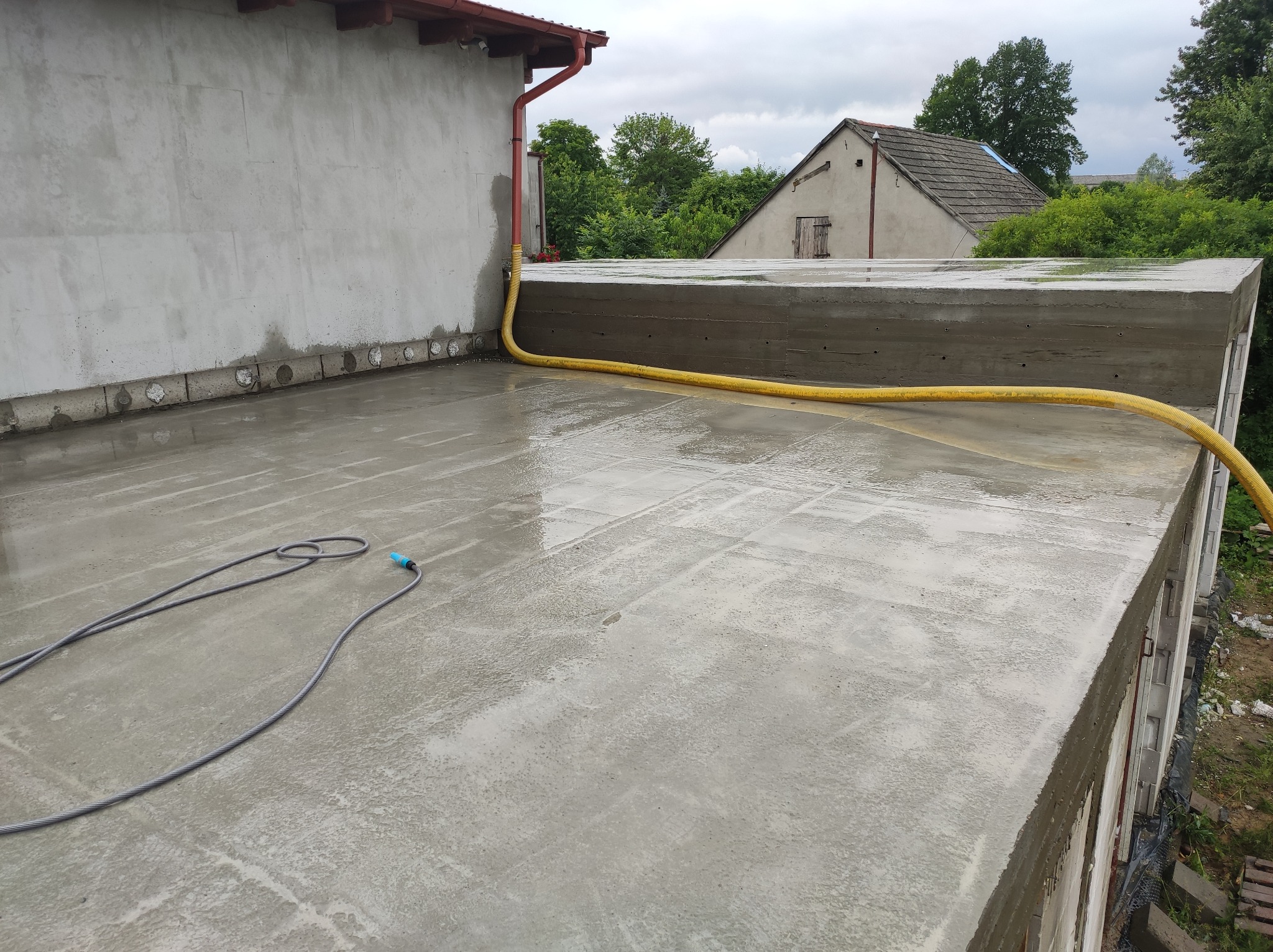 Świeżo wylany, mokry strop betonowy z widocznymi śladami po szalunku, z żółtym wężem do podlewania na powierzchni, w tle budynki i zieleń.