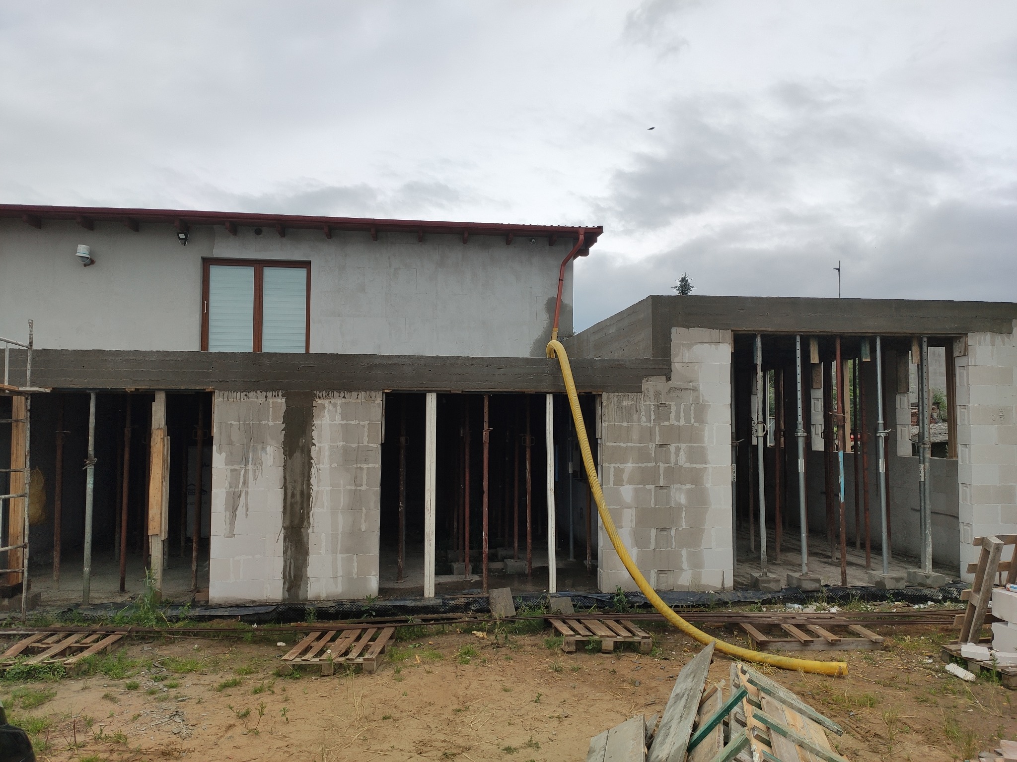 Budowa domu: surowy stan z widocznymi ścianami z bloczków betonowych, drewnianymi szalunkami i żółtym wężem doprowadzającym wodę, widok na dwukondygnacyjny budynek w trakcie wznoszenia.