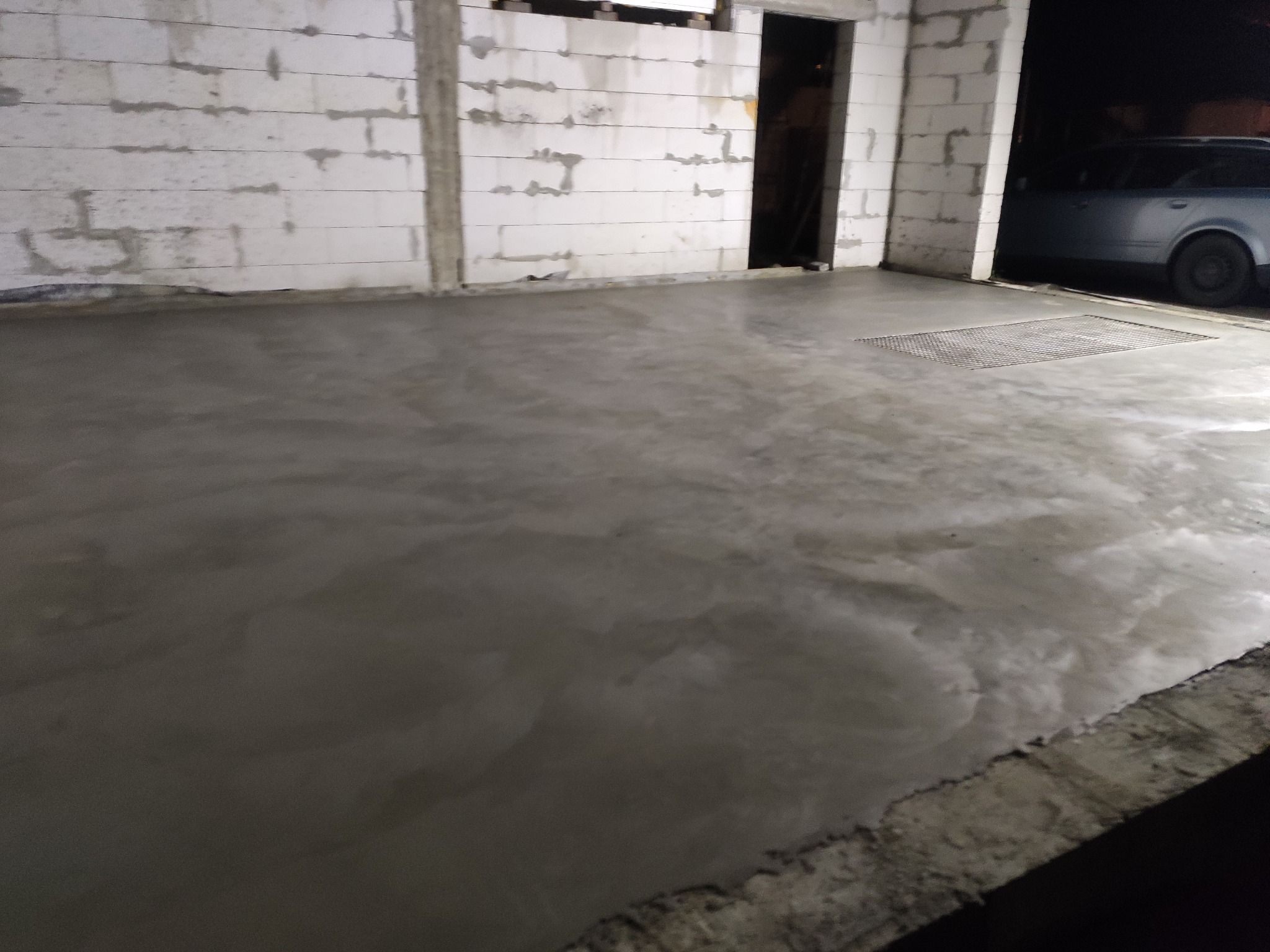 Świeżo wylana, gładka posadzka betonowa w surowym wnętrzu budynku z bloczków, widoczna kratka ściekowa i częściowo samochód na zewnątrz.