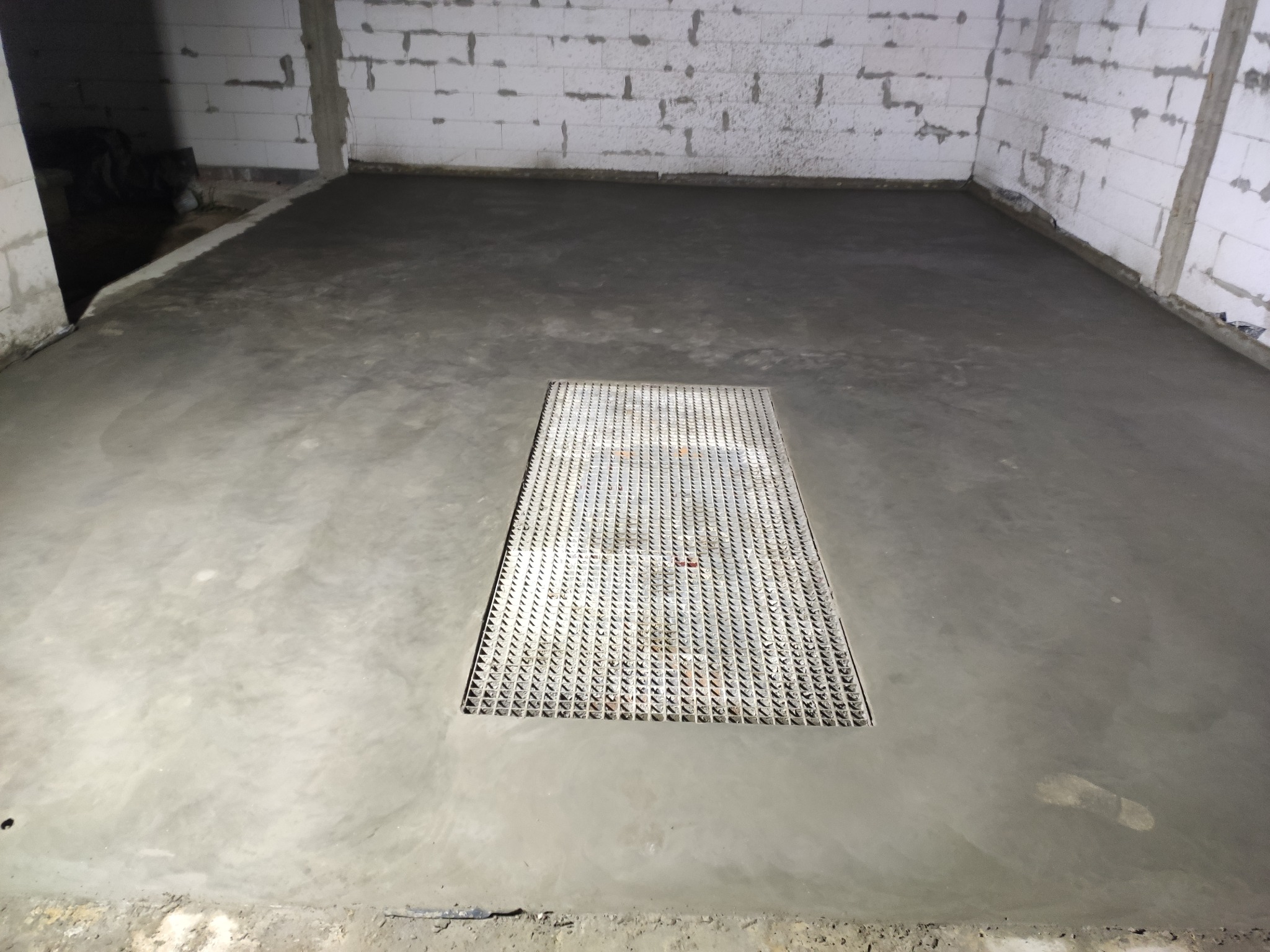 Świeżo wylana, gładka posadzka betonowa w surowym pomieszczeniu z pustaków, z metalową kratką ściekową pośrodku i odciskiem buta w rogu.