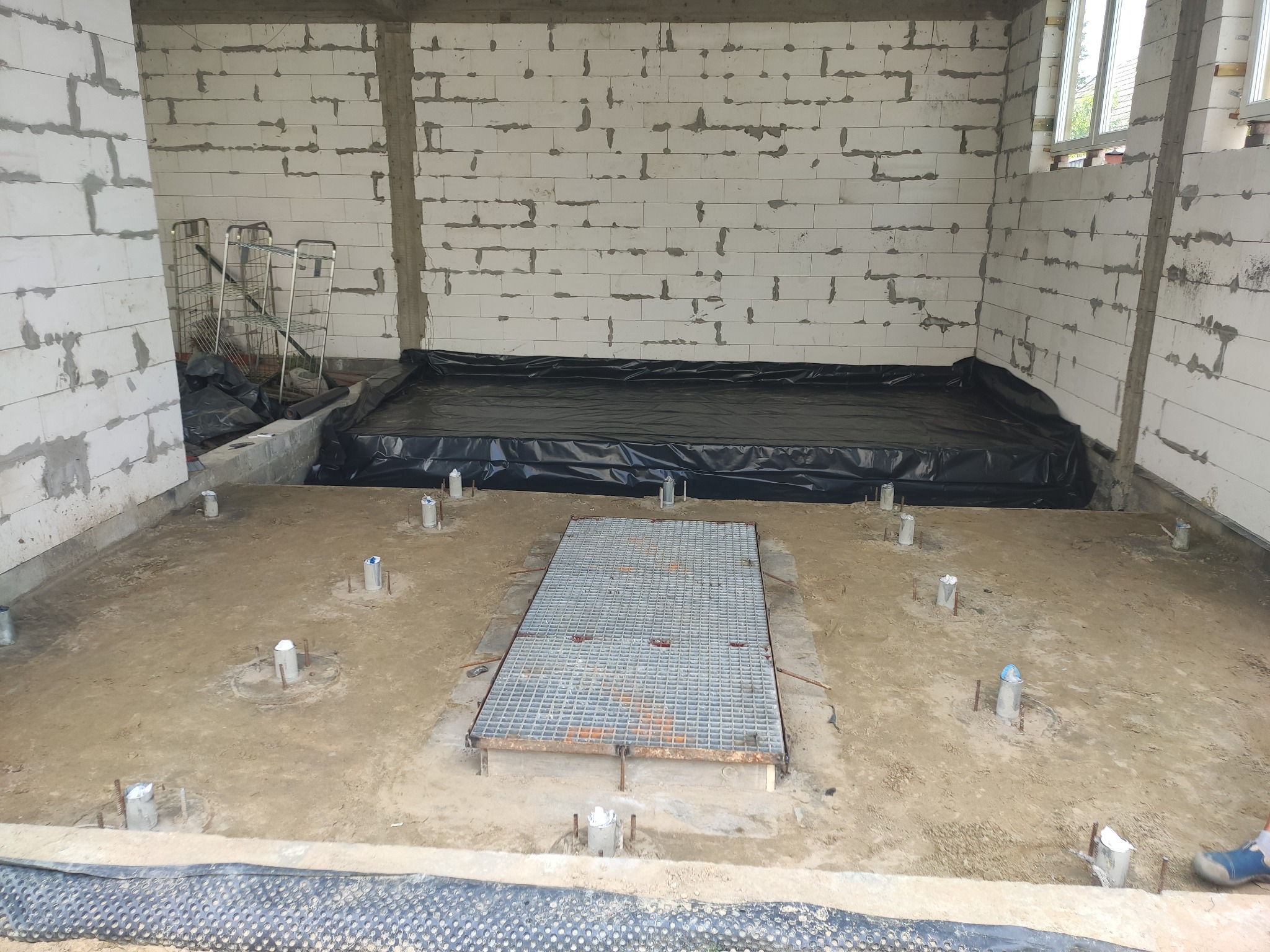 Wnętrze budynku w stanie surowym: ściany z białych bloczków, przygotowany teren pod wylewkę z wystającymi słupkami, czarna folia zabezpieczająca i metalowa krata w podłodze.