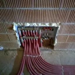 Rozdzielacz ogrzewania podłogowego zamontowany w ścianie z cegły, zabezpieczony pianką montażową, z widocznymi czerwonymi rurami rozprowadzającymi.