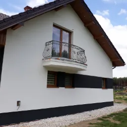 Nowoczesny dom jednorodzinny z białą elewacją, ozdobnym balkonem z metalową balustradą i ciemnymi akcentami w postaci pasów na ścianie i dachu, otoczony żwirem i zielenią.