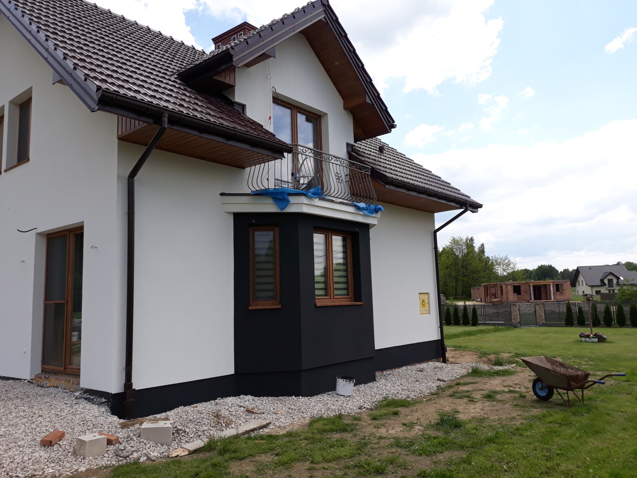 Dwupiętrowy dom po remoncie elewacji, z białymi ścianami i czarnym cokołem, widok z boku na ogród z taczką i placem budowy w tle.
