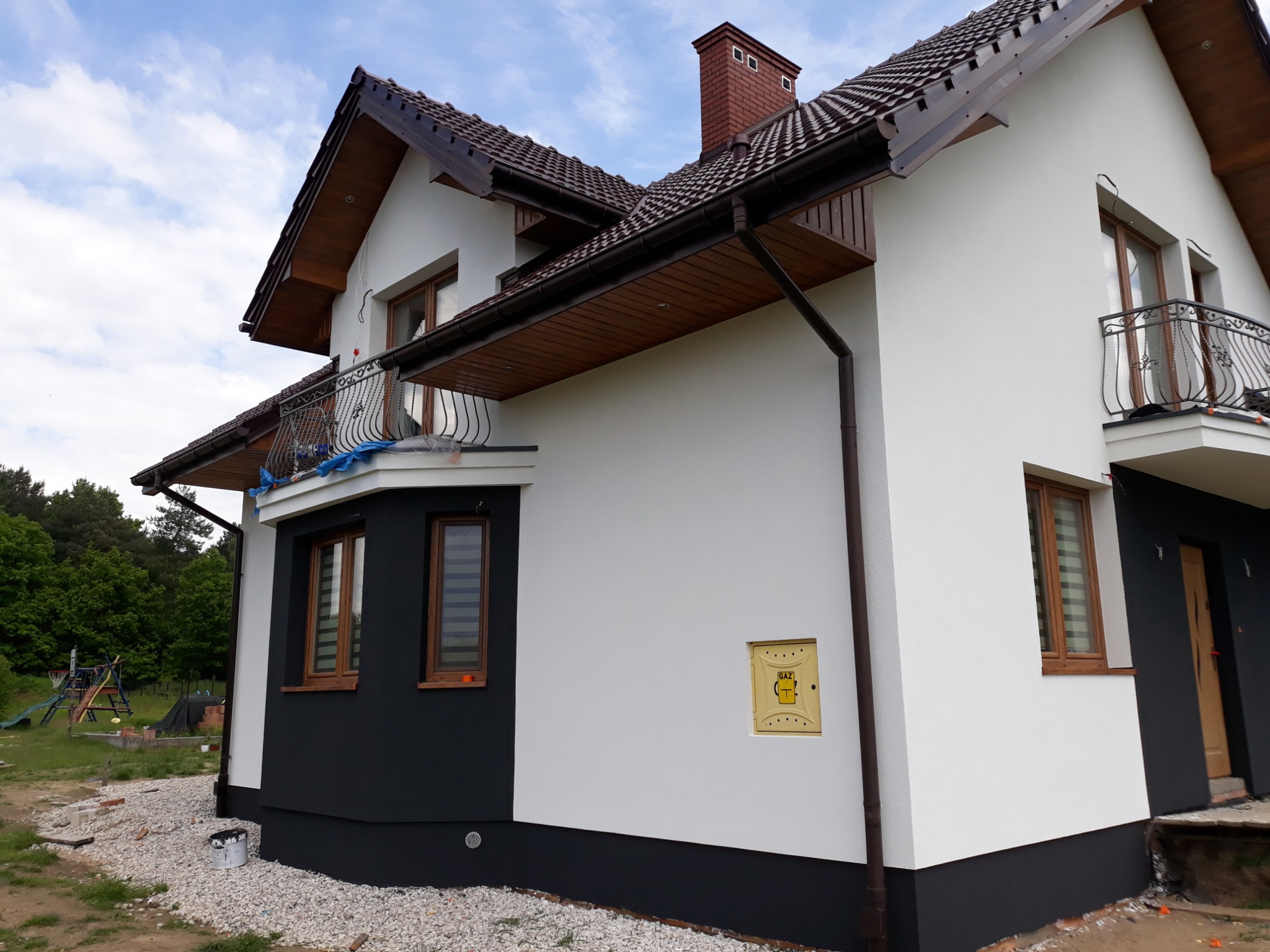 Dwukondygnacyjny dom z białą elewacją i ciemnobrązowymi akcentami, widok z boku na świeżo otynkowaną ścianę, z widoczną skrzynką gazową i fragmentem placu zabaw w tle.