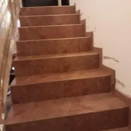 Schody wyłożone brązowymi płytkami ceramicznymi, prowadzące do ciemnych drzwi. Widoczne niedociągnięcia na ścianie wymagające wykończenia.