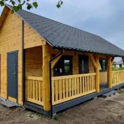 Nowoczesna altana z drewna o naturalnym odcieniu, z ciemnymi drzwiami i dachem, otoczona zielenią i drzewami.