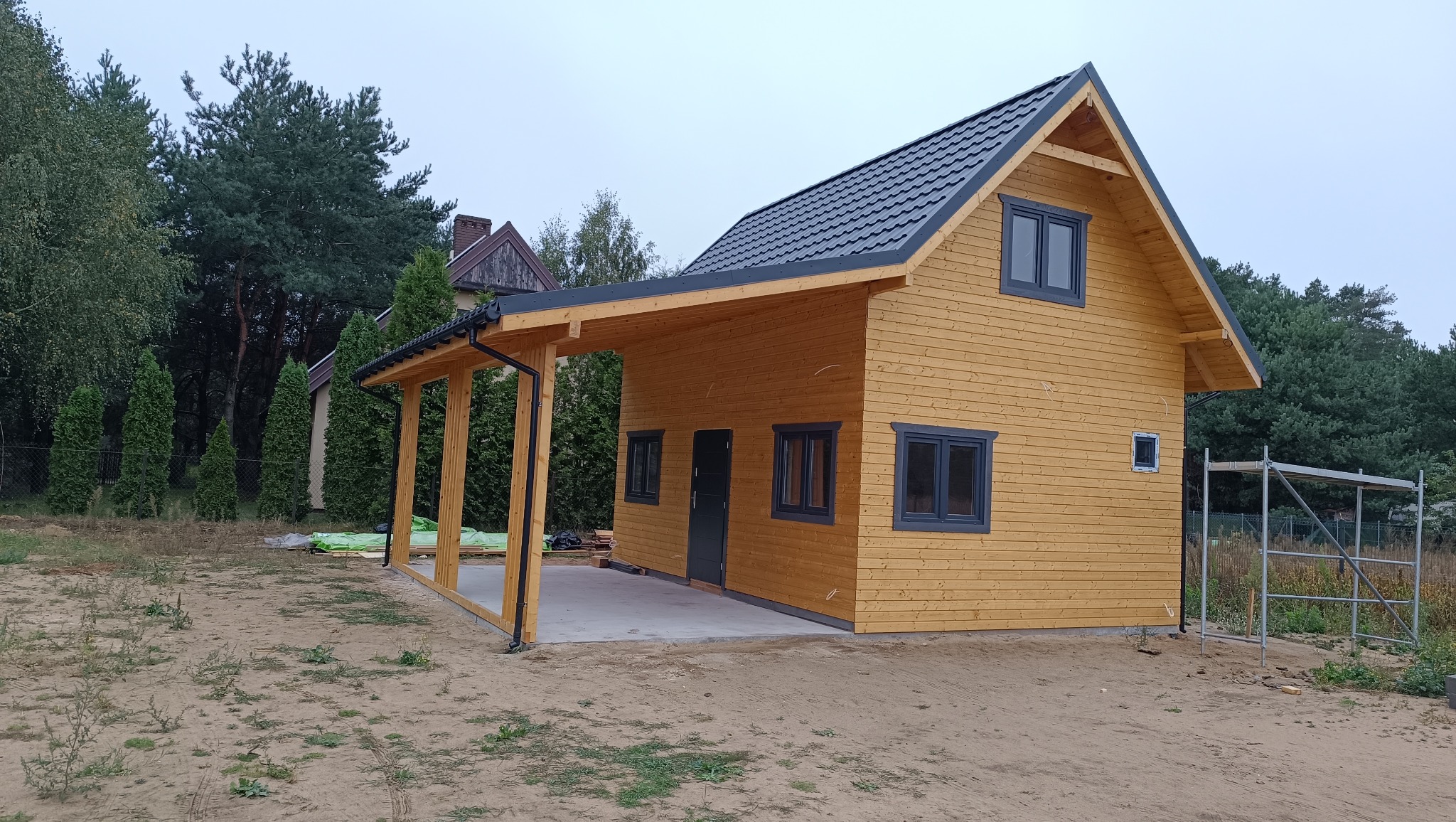 Nowoczesna, drewniana konstrukcja z zadaszeniem w trakcie budowy, widoczne rusztowanie i materiały budowlane w otoczeniu drzew.