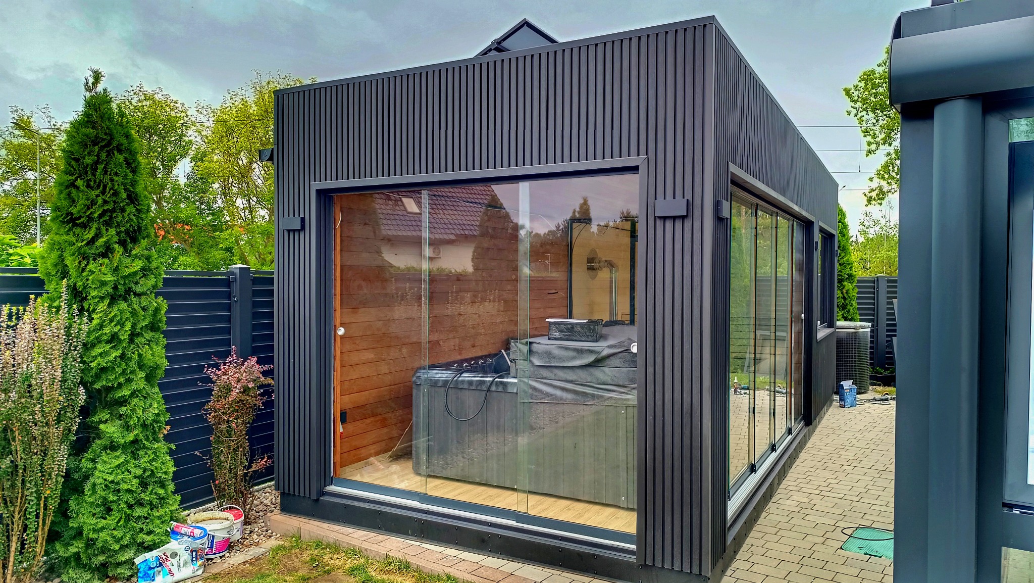 Nowoczesna, ciemnoszara altana z dużymi przeszkleniami i drewnianym wnętrzem, w której znajduje się jacuzzi przykryte pokrowcem, otoczona zielenią i ogrodzeniem.