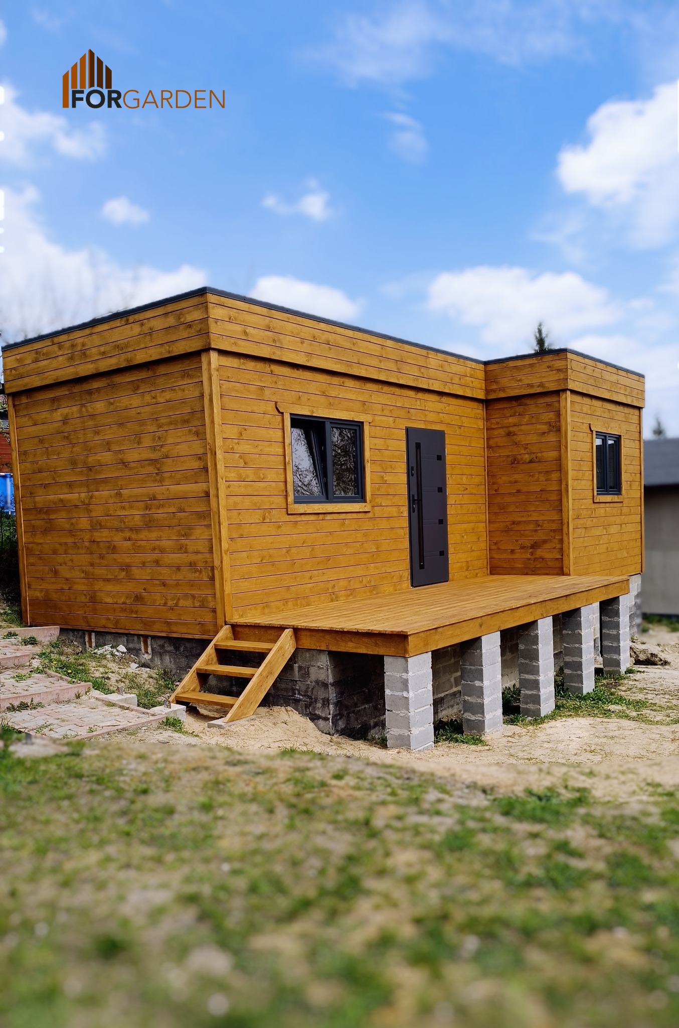 Nowoczesna, drewniana konstrukcja z płaskim dachem, podniesiona na betonowych fundamentach, z drewnianym tarasem i schodami, na tle błękitnego nieba z chmurami. Widoczne logo firmy 'FORGARDEN'.