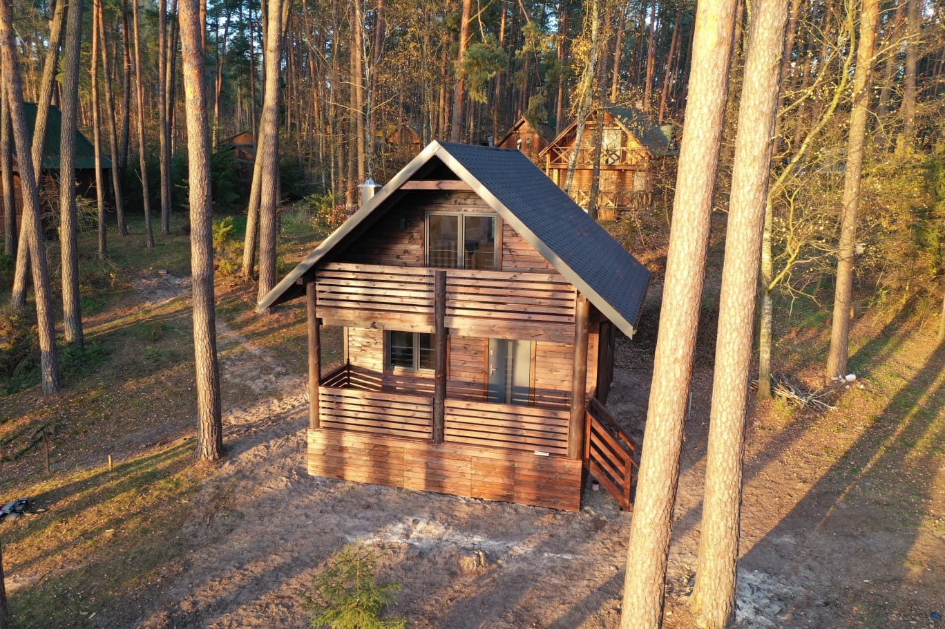 Drewniana konstrukcja w lesie, prawdopodobnie domek letniskowy, z balkonem, schodami i ciemnym dachem, otoczona wysokimi drzewami w promieniach zachodzącego słońca.
