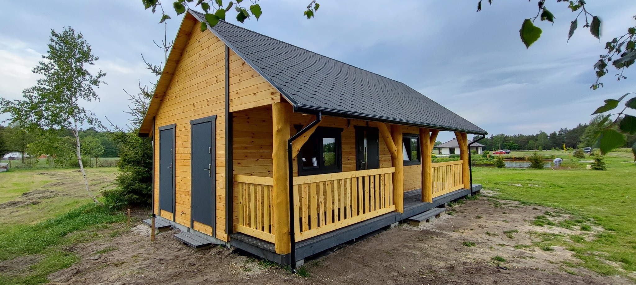 Nowoczesna altana z drewna o naturalnym odcieniu, z ciemnymi drzwiami i dachem, otoczona zielenią i drzewami.