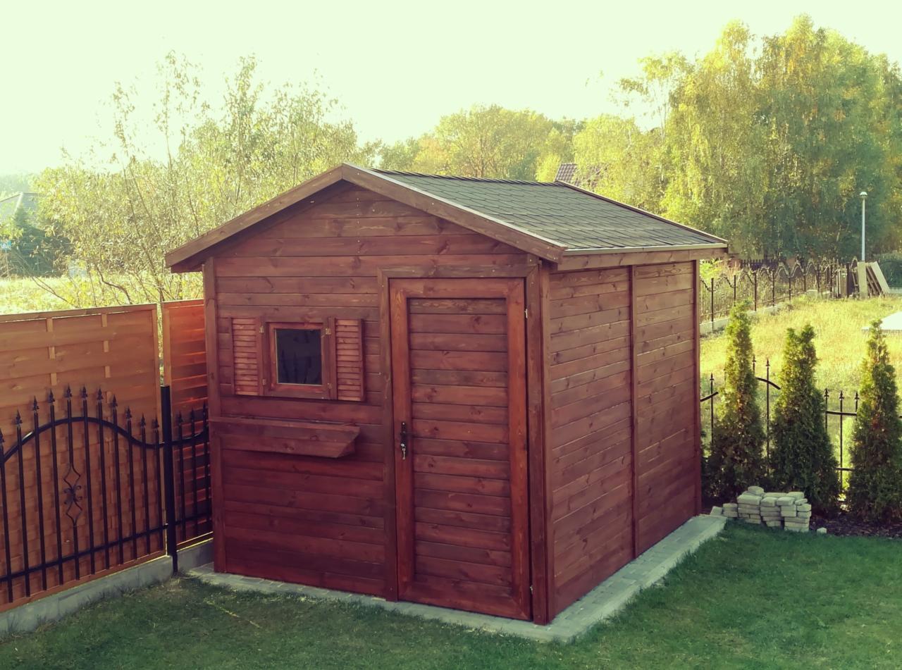 Drewniana altana ogrodowa w kolorze ciemnego brązu, z małym oknem z okiennicami i drzwiami, na tle zielonego trawnika i ogrodzenia z metalowymi elementami.