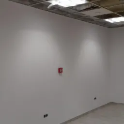 Wnętrze biura po malowaniu, widoczne gładkie, białe ściany, częściowo odsłonięty sufit podwieszany, czujnik przeciwpożarowy i gniazdka elektryczne.