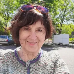 Uśmiechnięta kobieta w tweedowej marynarce i okularach na głowie, na tle drzew i zaparkowanych samochodów.