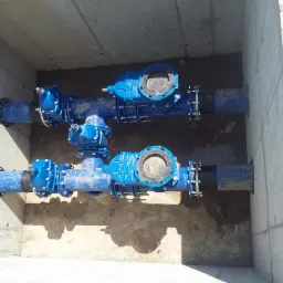 Dwa równoległe, niebieskie rurociągi z zaworami w betonowym wykopie, połączone czarnymi łącznikami, widoczne ślady ziemi na rurach.