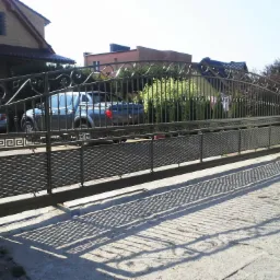 Przesuwna brama wjazdowa z metalu, zdobiona ornamentami, z widocznym samochodem osobowym w tle, oświetlona silnym światłem słonecznym rzucającym wyraźne cienie na podjazd.