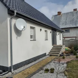 Świeżo pomalowana na biało elewacja domu z ciemnoszarą dachówką, widoczne detale wykończeniowe z kamienia elewacyjnego, rynny i satelita na ścianie, kostka brukowa prowadząca do wejścia.