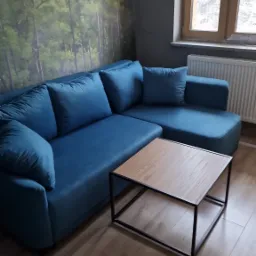 Turkusowy narożnik z poduszkami, stolik kawowy z drewnianym blatem na metalowej ramie, fototapeta z motywem leśnym i okno z grzejnikiem w tle.