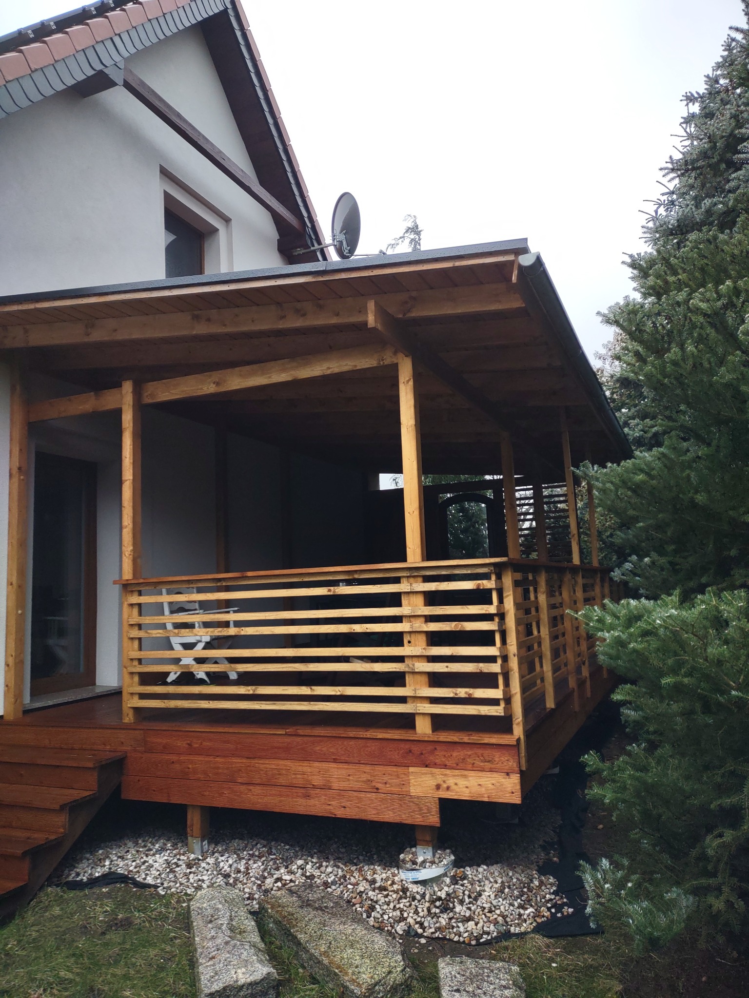 Drewniana altana z balustradą, przylegająca do domu z białą elewacją, widoczna antena satelitarna, otoczona zielenią i kamieniami.