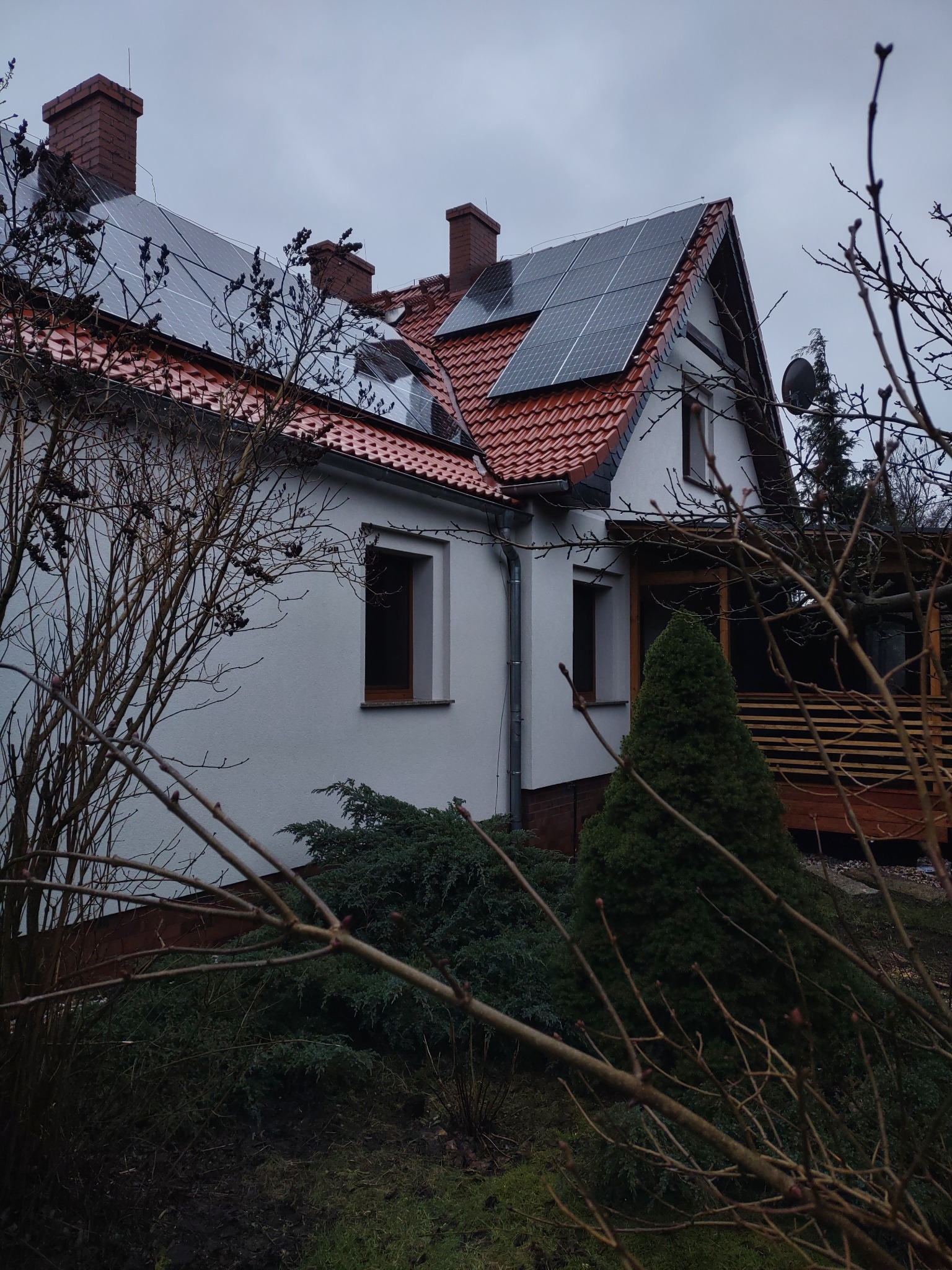 Dom jednorodzinny z białymi ścianami, czerwoną dachówką i panelami słonecznymi na dachu, widziany przez gałęzie drzewa.