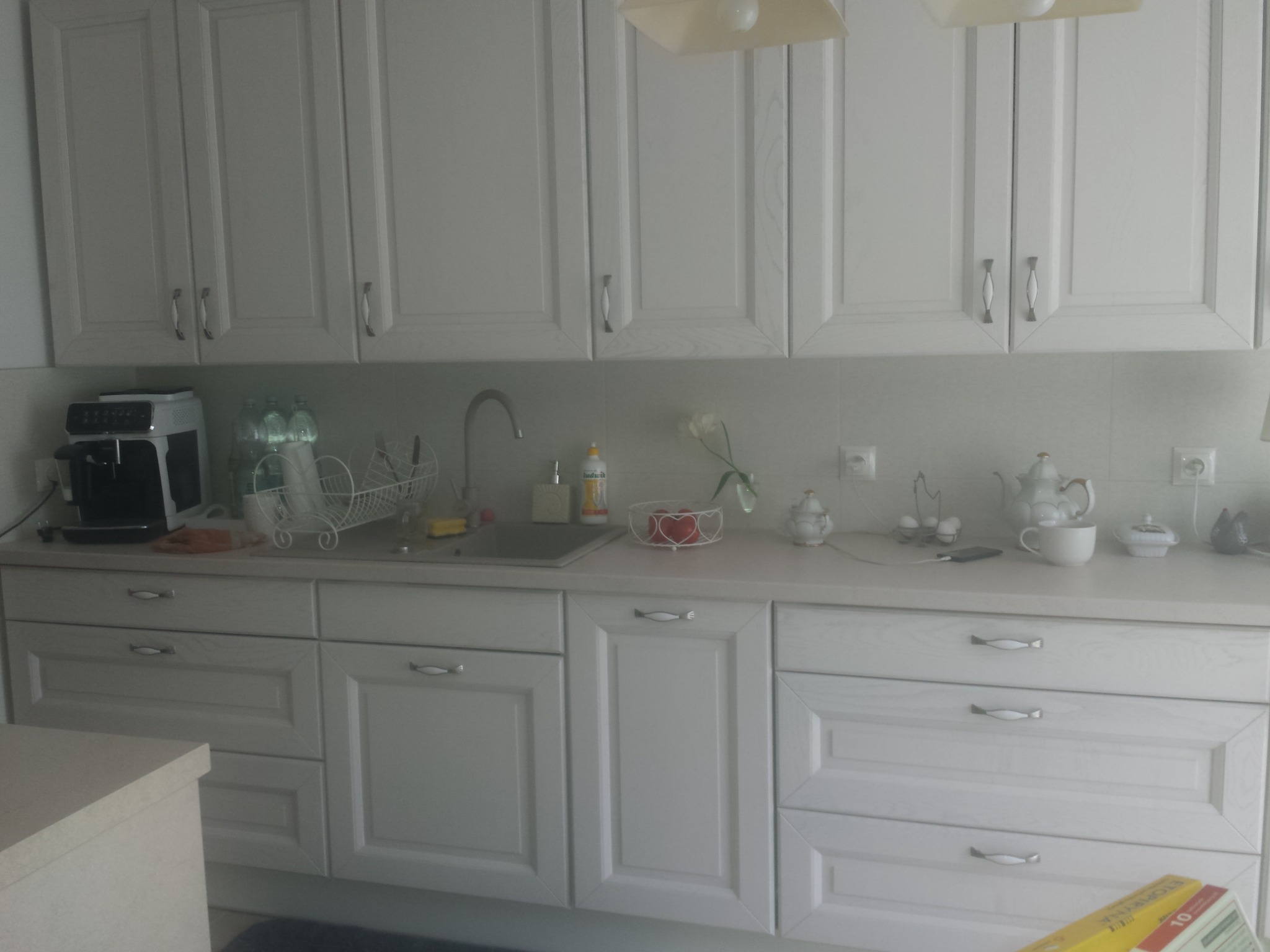 Jasna kuchnia z białymi, frezowanymi szafkami, srebrnymi uchwytami, blatem w odcieniu złamanej bieli, zlewozmywakiem i różnymi akcesoriami kuchennymi, takimi jak ekspres do kawy, naczynia i dekoracje.