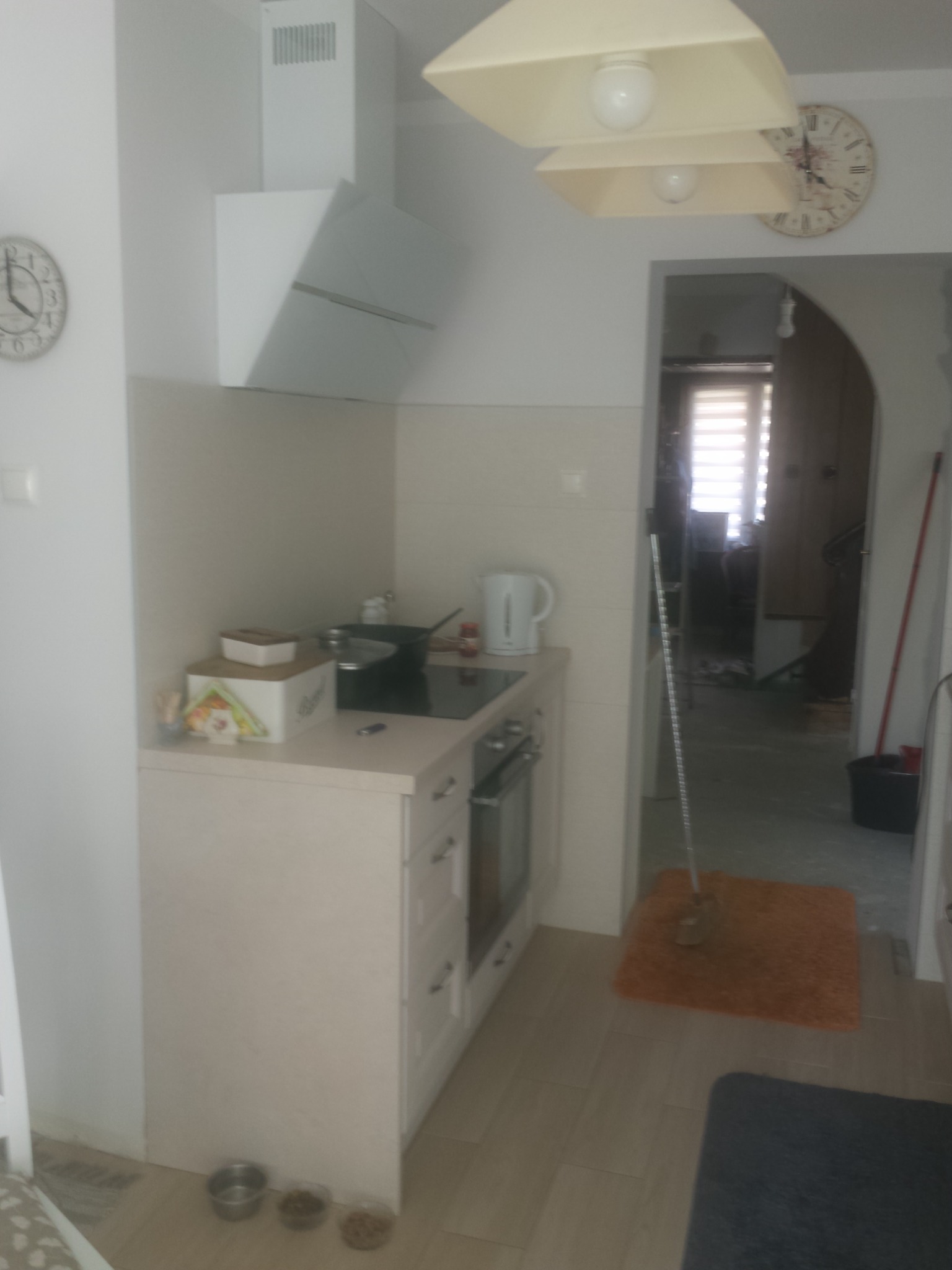 Jasna kuchnia z kremowymi szafkami, białym okapem i widocznym wejściem do drugiego pomieszczenia, w którym widać mop i niebieski dywanik.