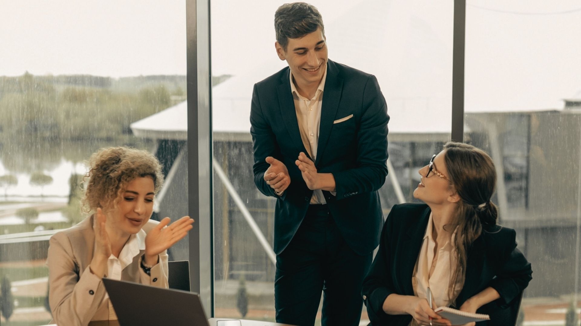 Trzy osoby w biurze: mężczyzna w garniturze klaszcze, a dwie kobiety przy biurku uśmiechają się i również klaszczą, widok za oknem na zewnątrz.