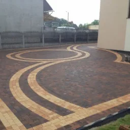 Ułożona kostka brukowa w odcieniach brązu i beżu tworząca wzór fal i łuków na podjeździe przed domem, widoczny fragment ogrodzenia i elewacji budynku.